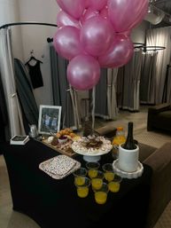 Shiny pink balloons floating over a party-ready spread of donuts, pastries, charcuterie, orange juice and wine set inside a boutique fitting room with curtained stalls and a hanging dress.