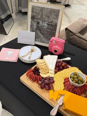 Birthday party table with a charcuterie board of cheese wedges, crackers, grapes and olives, stacked plates with gold forks, a pink instant camera, and a framed 'June' birthday sign.