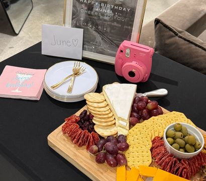 Birthday party table with a charcuterie board of cheese wedges, crackers, grapes and olives, stacked plates with gold forks, a pink instant camera, and a framed 'June' birthday sign.