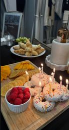 Festive birthday snack spread on a wooden board: sprinkled glazed donuts with lit candles, bowl of fresh raspberries, crackers and cheddar slices; plate of croissants and green grapes with a bottle of rosé in a marble cooler in the background.