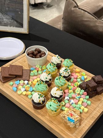 Wooden dessert board on a black tablecloth at an indoor party table featuring mini cupcakes with green and white frosting and sprinkles, chocolate squares, a ramekin of chocolate-covered almonds, colorful marshmallow cereal and brownie bites.