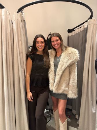 Two smiling women in a boutique fitting room trying on outfits — one in a beige faux-fur coat and white boots, the other in a black top and leggings.