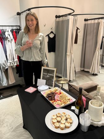 Smiling guest holding a small cup beside a birthday dessert spread with cupcakes, cookies and wine on a table inside a clothing boutique near fitting-room curtains and clothing racks.
