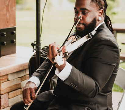 Musician in a black suit playing a white electric violin while seated under an outdoor pavilion at a green park-style venue