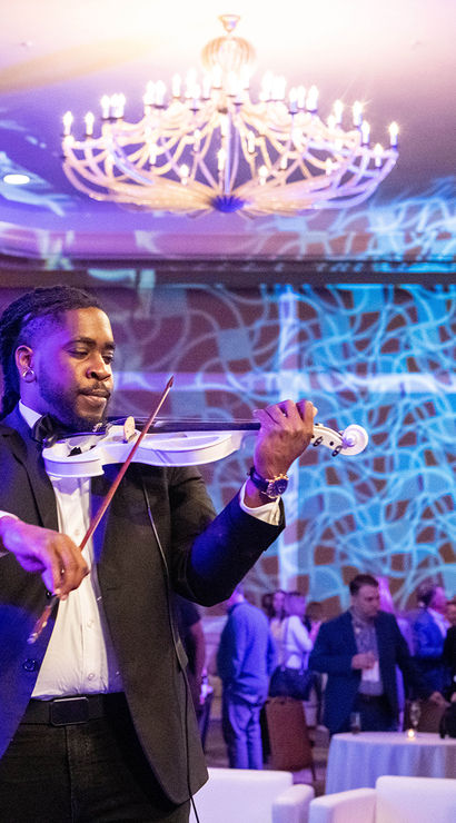 Violinist in a suit playing a white electric violin under purple-blue event lighting and chandelier in a ballroom, with guests mingling in the background - live performance at an indoor event.