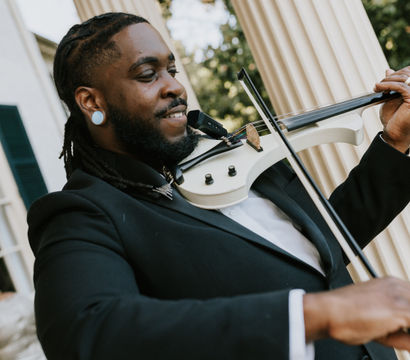 Electric violinist in a black suit with dreadlocks playing a white electric violin outdoors in front of tall classical columns — close-up of musician and instrument at a formal event.