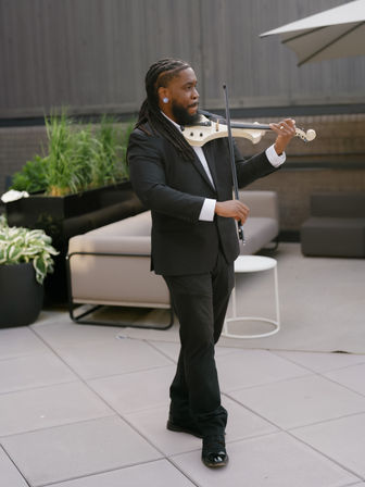 Violinist in a black suit with long dreadlocks playing a white electric violin on an outdoor urban patio with planters and lounge seating.