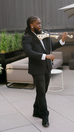 Violinist in a black suit with long dreadlocks playing a white electric violin on an outdoor urban patio with planters and lounge seating.