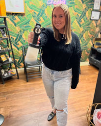 Smiling tanning technician holding a spray-tan gun toward the camera in a bright tanning salon with tropical leaf wallpaper and wood floors.