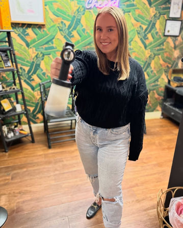 Smiling young woman in a bright beauty studio with tropical leaf wallpaper and wood floors, wearing a black sweater and ripped light jeans, holding a spray bottle toward the camera.