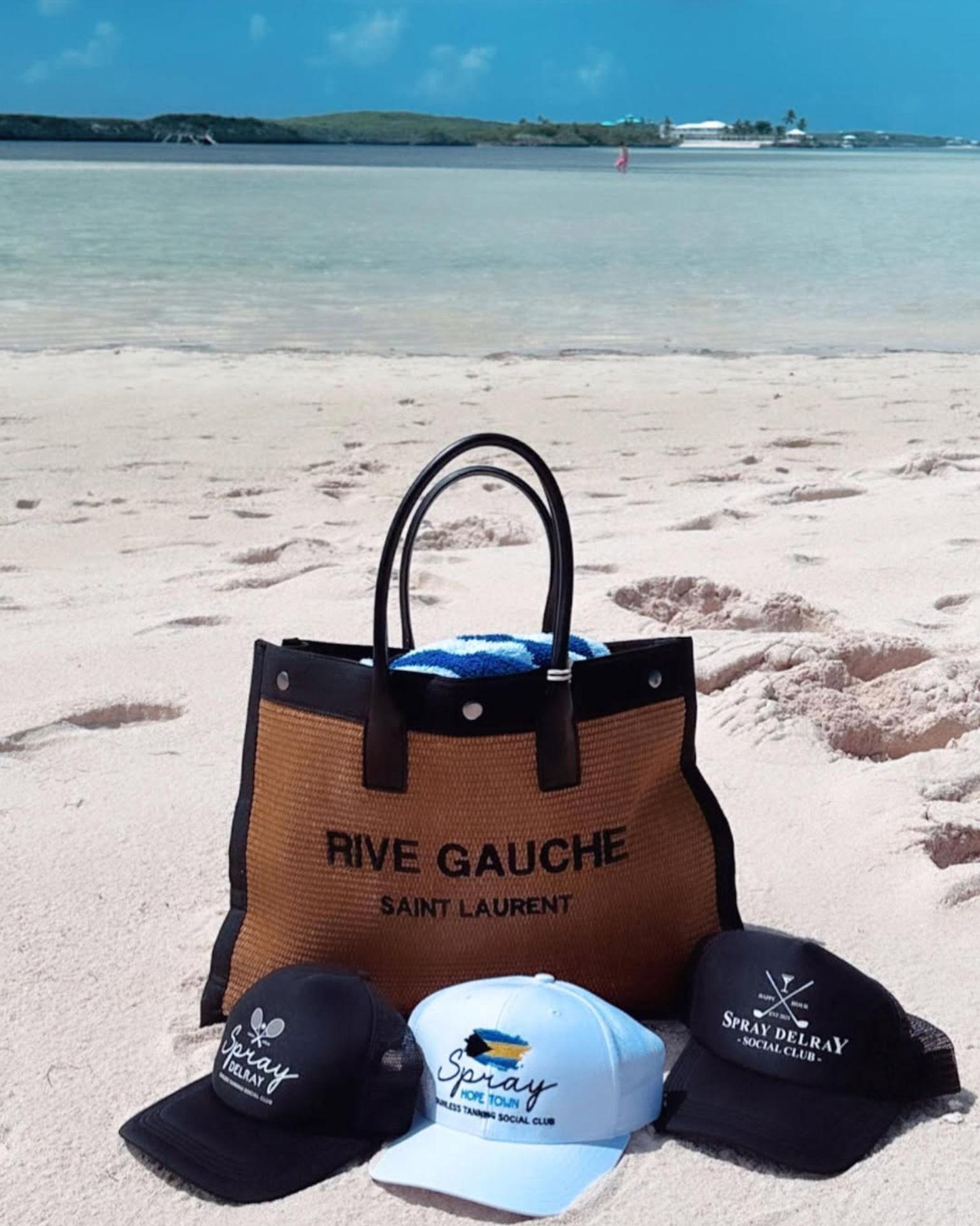Beach-ready woven tote with a blue towel and three baseball caps resting on white sand, turquoise shallow water and a low island on the horizon under a bright blue sky.