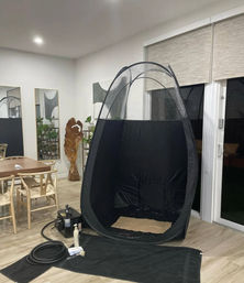 Portable black pop-up spray-tan tent with spray machine, hose and solution set up in a modern dining room near sliding glass doors