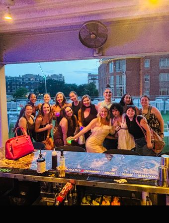 Group of friends celebrating on an outdoor rooftop bar patio at dusk, posing behind the bar with colorful drinks, a pink handbag on the counter, string lights and city buildings in the background.
