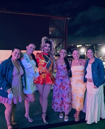 Smiling group of six women and a drag performer posing on a city rooftop at night, wearing colorful dresses and a bride-to-be sash for a bachelorette celebration.