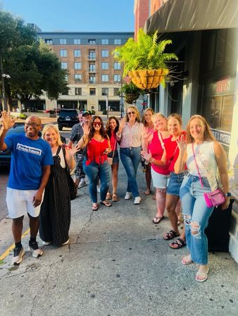 Cheerful group of friends waving on a sunny downtown sidewalk during a summer outing, wearing casual clothes with hanging fern planters and brick storefronts in the background.