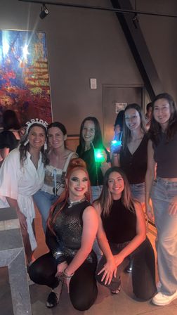 Smiling group of seven women posing in a dim lounge: two kneeling in front (one in a sparkly stage outfit with dramatic makeup), five standing behind (one wearing a 'BRIDE' headband), a guest holding a glowing green drink and colorful abstract artwork on the wall.