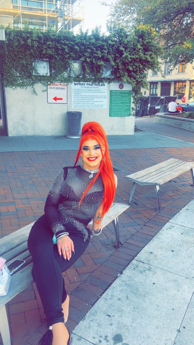 Smiling person with vivid red ponytail and dramatic makeup in a black rhinestone bodysuit, sitting on a bench in a downtown brick‑paved plaza with an ivy-covered wall and informational signs in the background.
