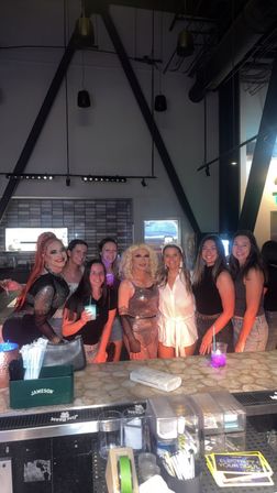 Friends and drag performers posing for a group photo at a lively bar with cocktails beneath industrial A-frame beams — fun nightlife celebration.