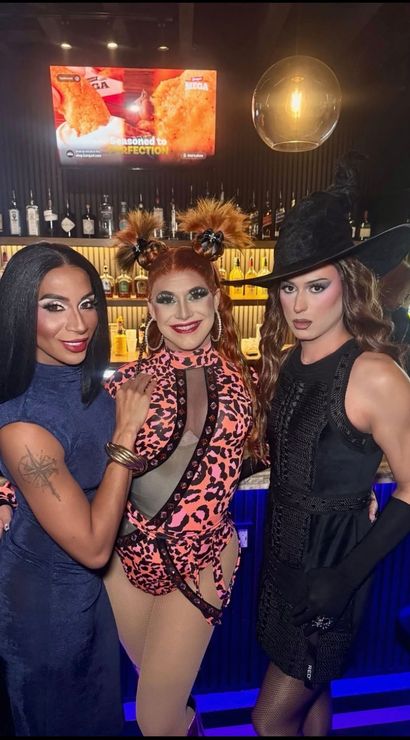 Three drag performers pose at a neon-lit nightclub bar — glam makeup and bold costumes: sleek navy dress, pink leopard bodysuit with playful space-bun hair, and a black witch-style outfit with tall hat, lively nightlife scene.