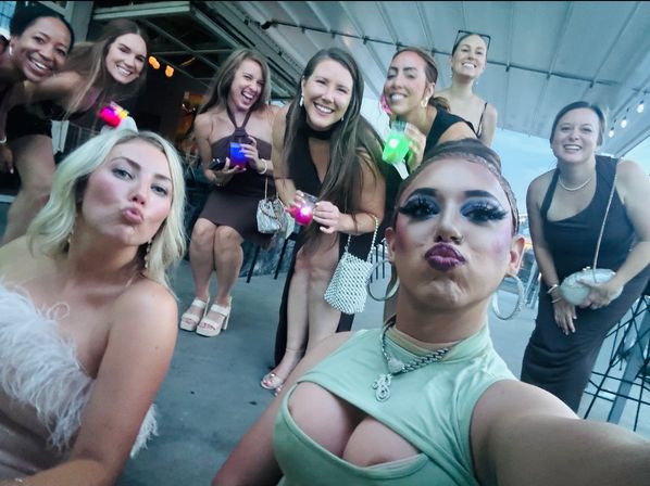 Rooftop bar group selfie of friends in evening attire, posing and puckering up with glowing cocktail cups on an outdoor terrace at dusk
