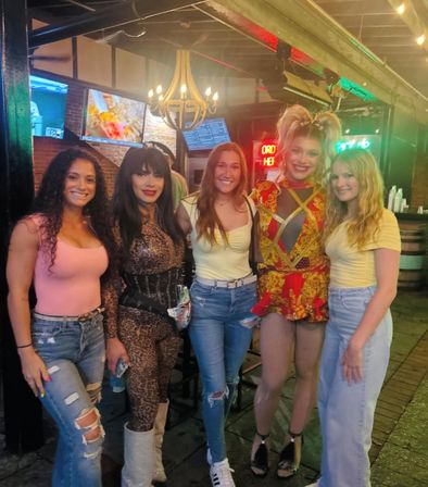 Nightlife bar group photo of five people smiling on a lively patio with neon signs and a chandelier; outfits include a leopard-print bodysuit with sparkly boots, a red-and-gold stage costume, and casual tops with ripped jeans.