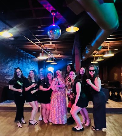 Group of women celebrating a bachelorette party on a dance floor in an urban lounge — bride in a sparkling gown with friends holding cocktails under a disco ball, exposed brick wall and colored ceiling lights.