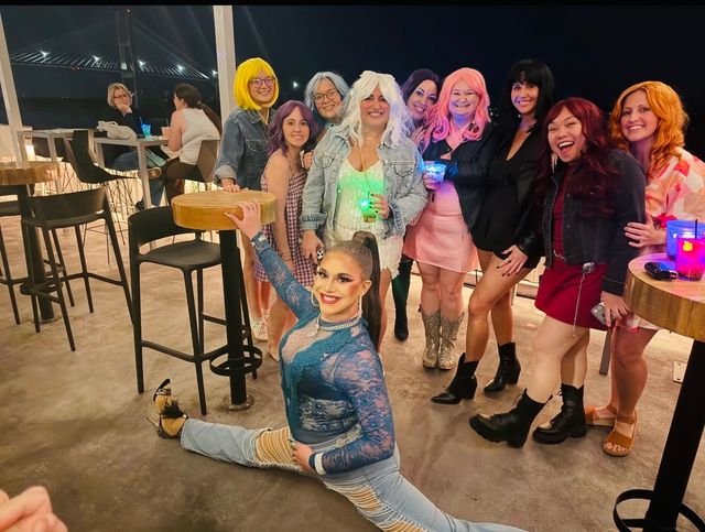 Performer doing a full split on a rooftop bar deck at night as a lively group of people in colorful wigs and outfits pose behind her with glowing drinks, illuminated suspension bridge and waterfront skyline in the background.