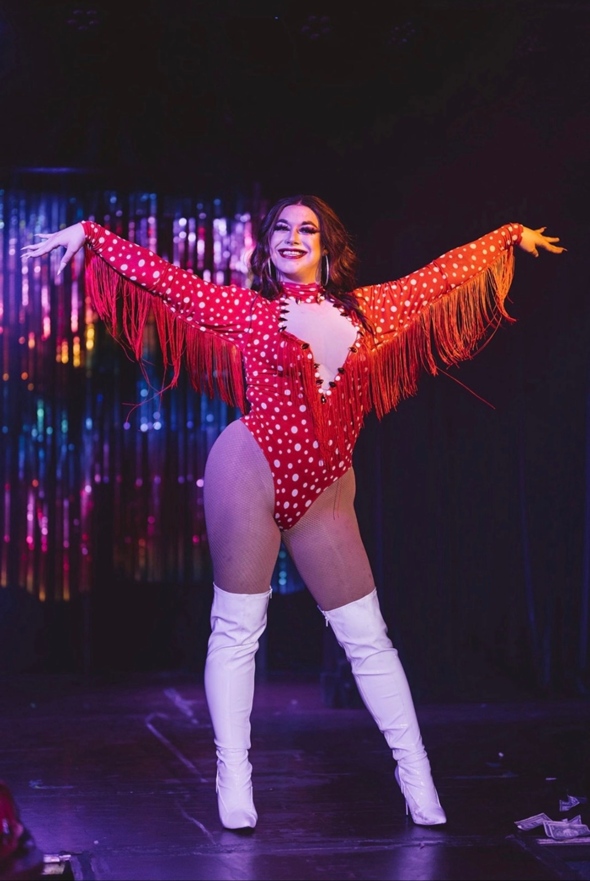 Drag performer on a neon-lit cabaret stage wearing a red polka-dot fringed bodysuit and white thigh-high boots, smiling with arms outstretched in front of a shimmering tinsel backdrop.