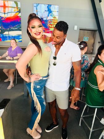 Smiling drag performer in a green top and fringe jeans poses with a playful friend at a lively indoor drag brunch, colorful abstract artwork and patrons in the background.