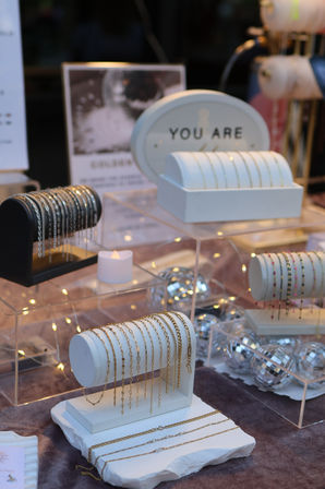 Close-up of a boutique jewelry display with delicate gold chains and bracelets draped over white cylindrical stands, mirrored disco balls and warm fairy lights creating a cozy window-stall ambiance.