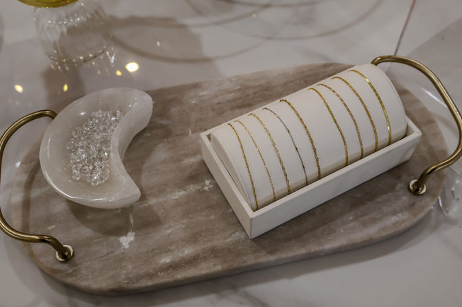 Delicate gold and silver chain bracelets arranged on a white cuff display atop a stone tray with brass handles, next to a crescent dish of sparkling clear beads — boutique jewelry display.