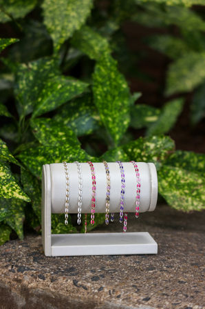 Close-up of colorful delicate chain bracelets in gold, silver, pink and purple draped on a white jewelry display stand on a stone ledge with green variegated foliage garden backdrop