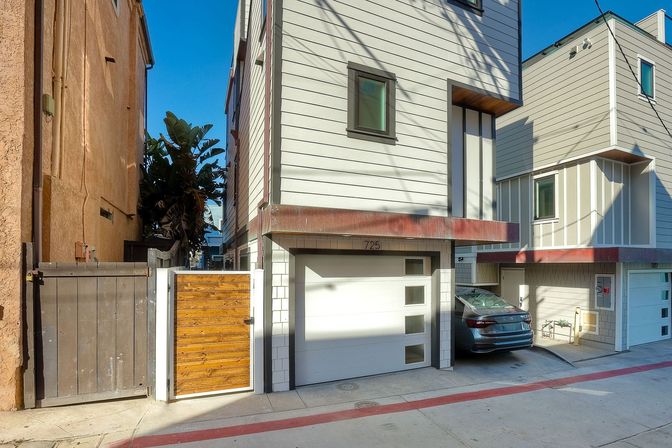 Sunlit narrow modern townhouse with white garage door, wooden side gate, parked sedan and palm tree in a bright urban alley