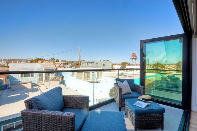 Sunny rooftop balcony with modern wicker lounge chairs and ottoman with blue cushions, straw hat and magazine on the ottoman, glass railing and low-rise neighborhood view under a clear blue sky
