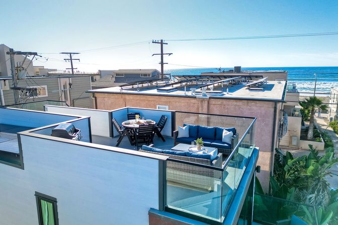 Sunlit ocean-view rooftop patio in a beachside neighborhood with glass railings, blue-cushioned wicker sofa, round dining table set for four, and a gas grill overlooking nearby buildings and the waves.