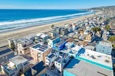 Aerial view of a sunlit coastal neighborhood with vibrant beachfront homes and modern condos lining a wide sandy beach, waves rolling in and a distant pier under a clear blue sky.