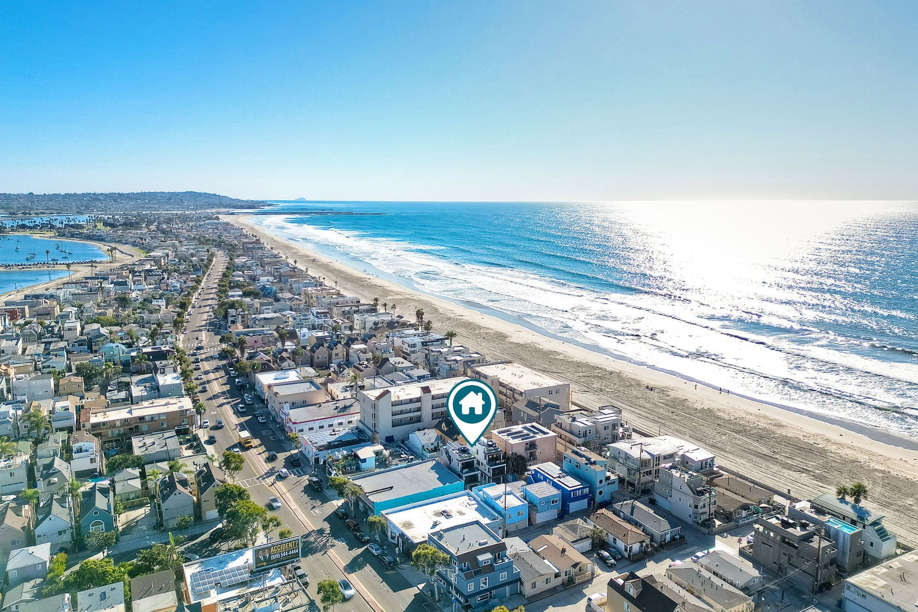 Aerial view of a sunlit oceanfront neighborhood with rows of beachfront homes along a long sandy shoreline, sparkling blue waves, and a map-pin icon marking a house.