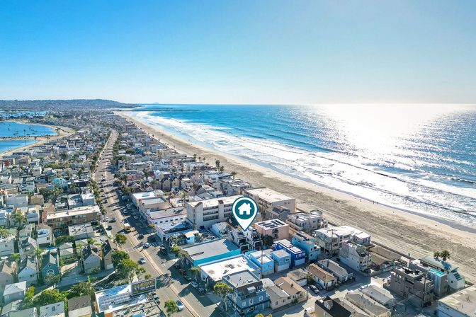 Aerial view of a sunlit oceanfront neighborhood with rows of beachfront homes along a long sandy shoreline, sparkling blue waves, and a map-pin icon marking a house.