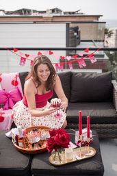 Smiling woman in a red top on an urban rooftop balcony holding a dessert glass at a pink-and-red Valentine's-themed picnic with roses, candles, and snacks.