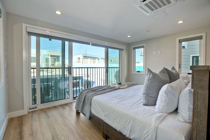 Bright modern bedroom with white bedding and gray throw pillows, sliding glass doors opening to a balcony, hardwood floors, and views of nearby buildings under a blue sky.