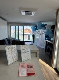 Sunlit seaside apartment in San Diego decorated for a coastal bachelorette: blue and white balloon arch reading 'Last Toast on the Coast', event signs on a marble island, sliding glass doors to a balcony with ocean-view homes.