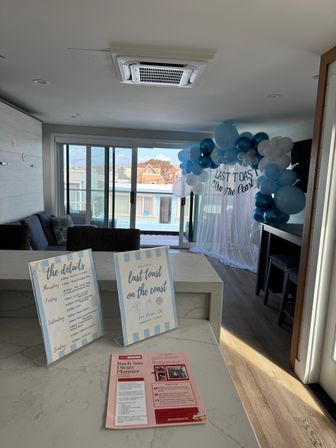 Sunlit seaside apartment in San Diego decorated for a coastal bachelorette: blue and white balloon arch reading 'Last Toast on the Coast', event signs on a marble island, sliding glass doors to a balcony with ocean-view homes.