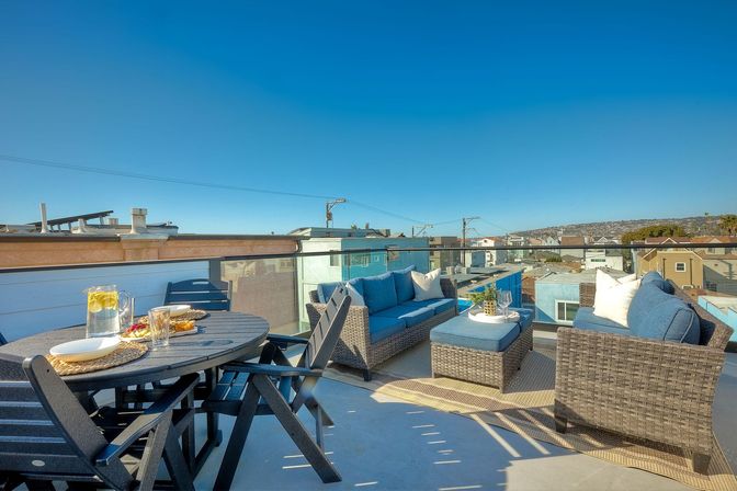 Sunny rooftop deck with a round dining table set with lemon water and snacks, navy-cushioned wicker sofa, chairs and ottoman, glass railing and clear blue sky over a coastal neighborhood.