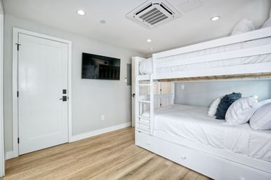 Bright modern coastal-style bedroom with white wooden bunk beds, crisp white linens and decorative pillows, wall-mounted flat-screen TV, light wood flooring and recessed ceiling lights.