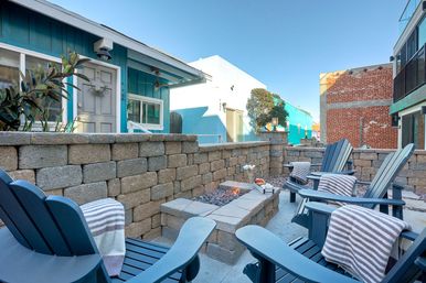 Inviting coastal courtyard with blue Adirondack chairs and striped throws gathered around a rectangular stone fire pit, stacked stone walls and a teal beach cottage under a clear blue sky.