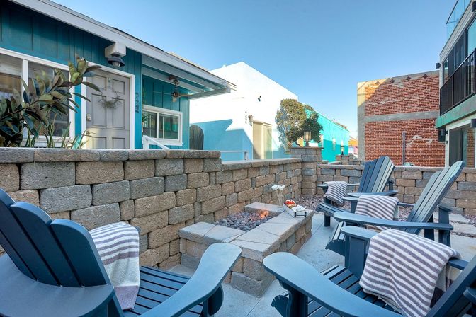 Inviting coastal courtyard with blue Adirondack chairs and striped throws gathered around a rectangular stone fire pit, stacked stone walls and a teal beach cottage under a clear blue sky.
