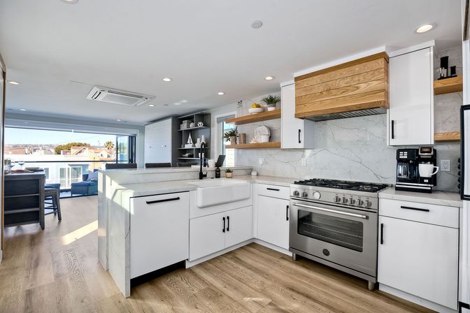 Bright modern open-concept coastal kitchen with marble countertops, farmhouse sink, stainless steel gas range, wooden hood, open shelving, light wood floors and sliding glass doors to a sunlit balcony with ocean view.
