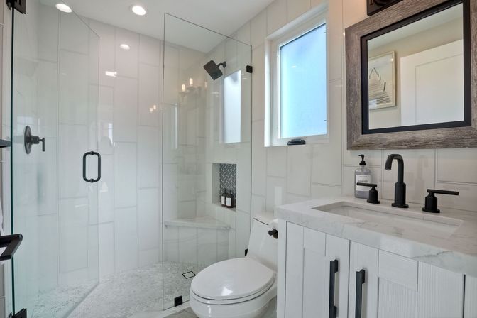 Bright modern white bathroom with frameless glass walk-in shower, pebble-tile floor, built-in shower niche, black matte fixtures, marble vanity and wood-framed mirror under a frosted window.