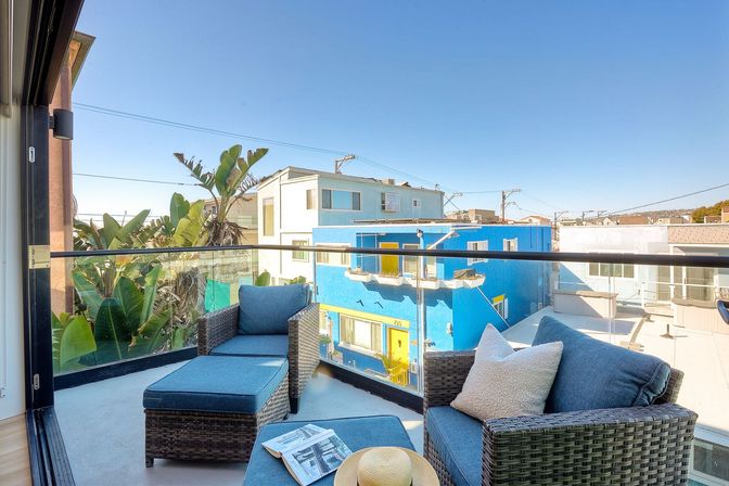 Sunny urban balcony with wicker lounge chairs and ottoman, blue cushions, straw sun hat and magazine on the ottoman, glass railing overlooking palm trees and a vibrant blue building under a clear sky.