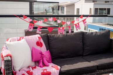Urban rooftop balcony sofa decorated for Valentine’s Day with pink and red heart garlands spelling BE MINE, a heart-print pink throw and a bright pink bow cushion.